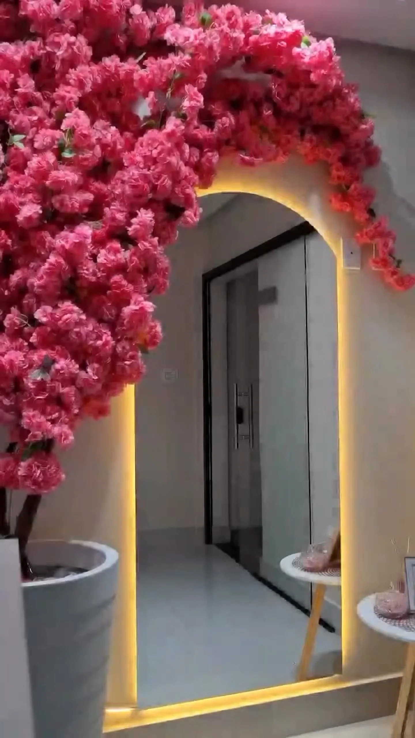 Espelho em arco iluminado por trás com luz âmbar, sob uma cascata de flores cor-de-rosa, na entrada do Studio Zoe