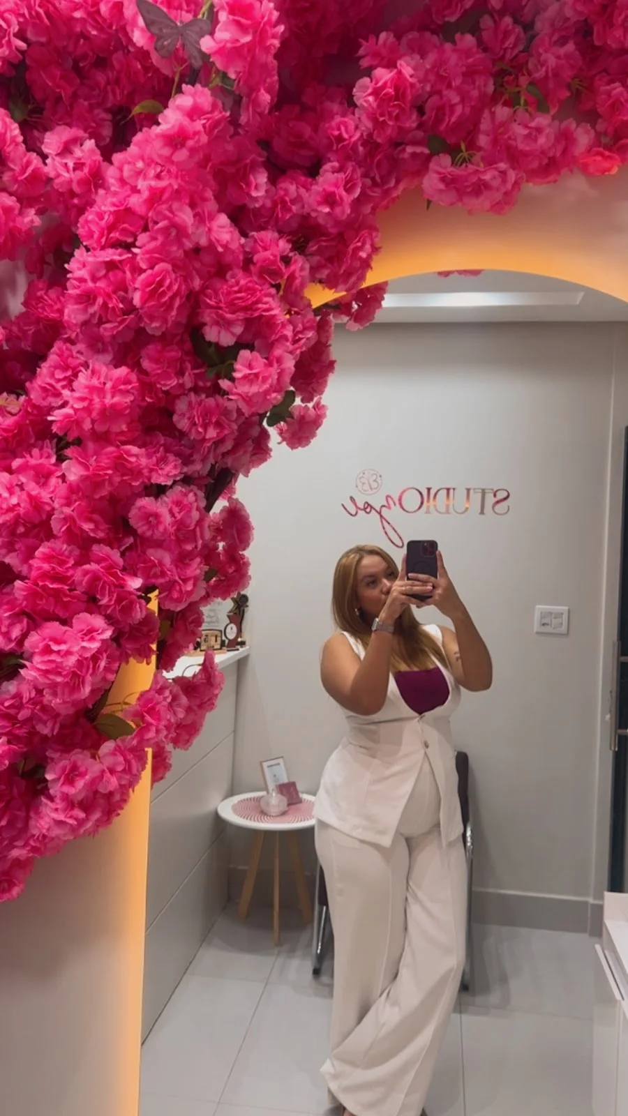Cynthia, de conjunto branco, fotografando-se no espelho em arco da entrada, com as flores cor-de-rosa e o letreiro Studio Zoe refletido